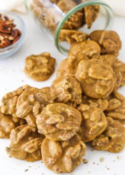 Southern Pecan Pralines poured out of a glass jar on it's side onto a white table with a bowl of pecans in the background