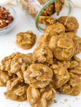 Southern Pecan Pralines poured out of a glass jar on it's side onto a white table with a bowl of pecans in the background