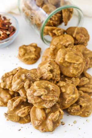 Southern Pecan Pralines poured out of a glass jar on it's side onto a white table with a bowl of pecans in the background