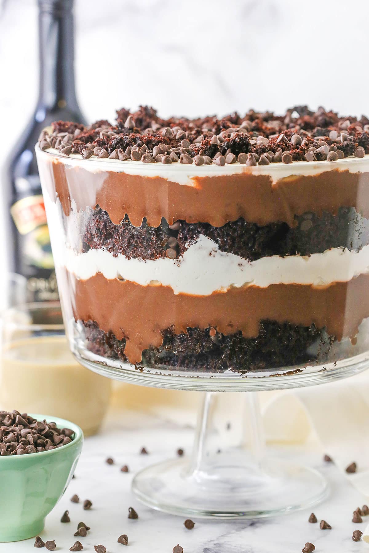 This Boozy Baileys Chocolate Trifle is made with layers of moist chocolate cake, Baileys whipped cream, Baileys chocolate pudding and chocolate chips! A tasty dessert that's so easy to make and great for a crowd! Side view of a full Boozy Baileys Chocolate Trifle in a glass trifle stand topped with chocolate chips