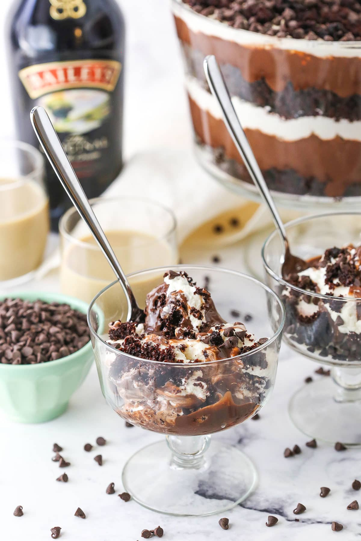 Two individual servings of Boozy Baileys Chocolate Trifle in glass bowls with spoons with chocolate chips, Baileys and the trifle in the background