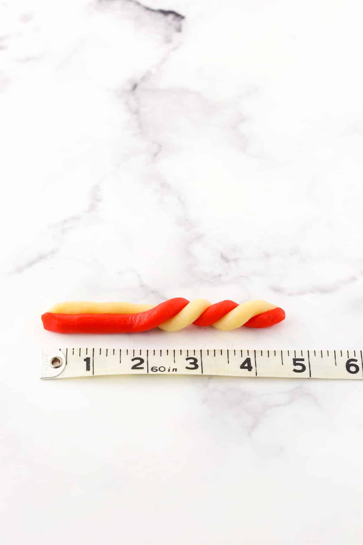 A step in making Red and white Candy Cane Cookies showing the twisting red and white batter to shape the candy cane next to a tape measure showing the scale and size