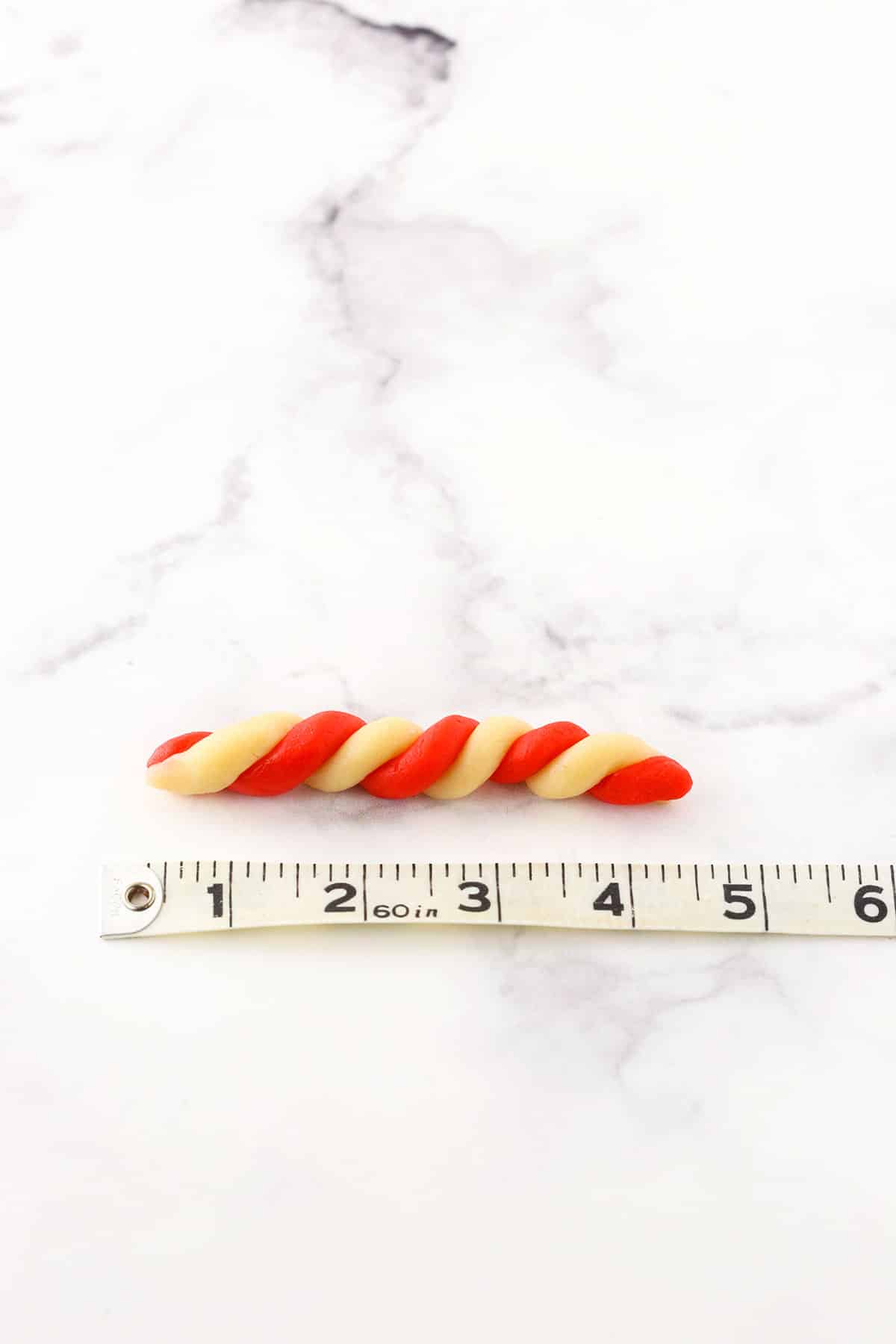 A step in making Red and white Candy Cane Cookies showing the twisting red and white batter to shape the candy cane next to a tape measure showing the scale and size