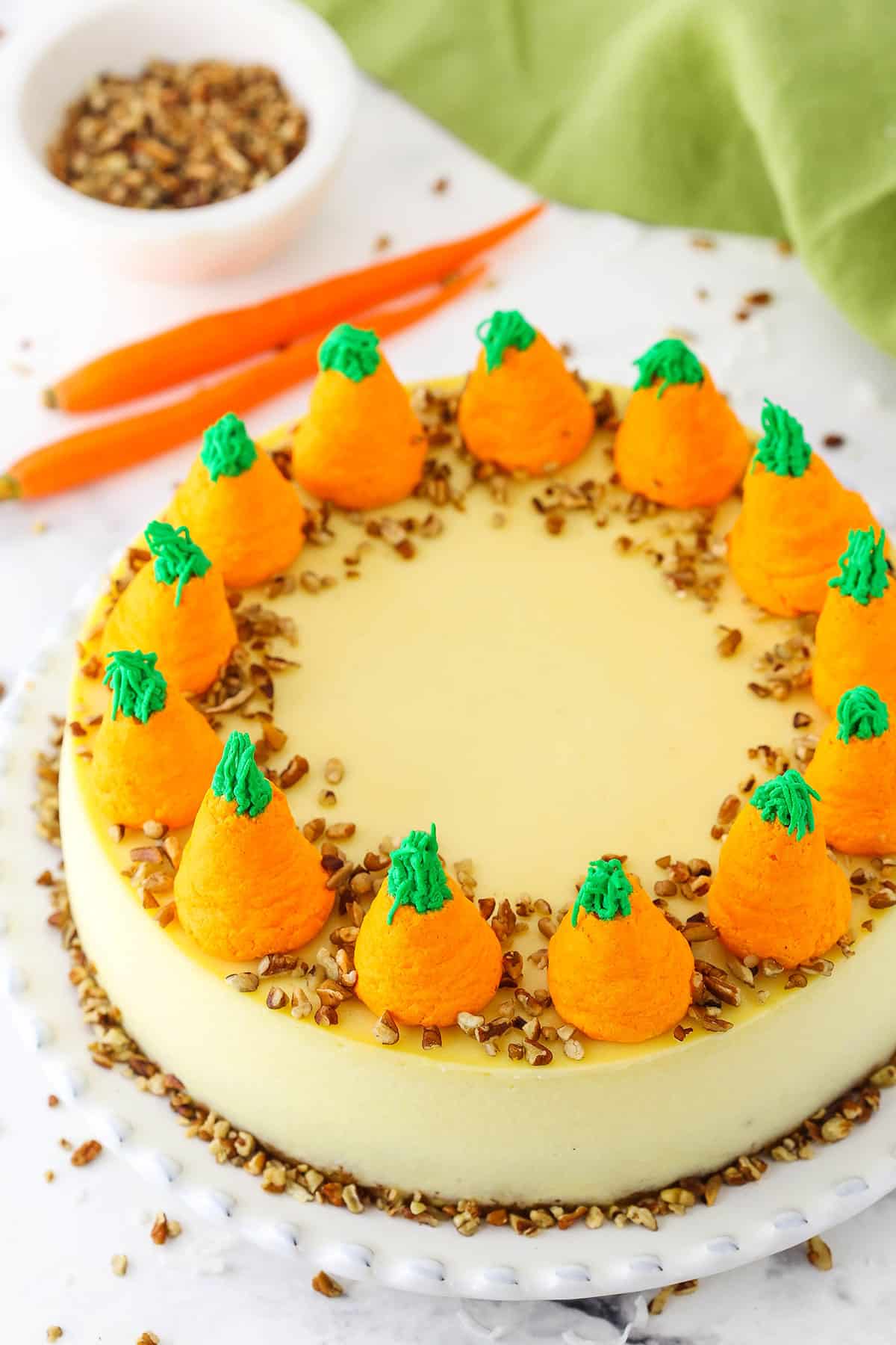 Overhead view of a full Carrot Cake Cheesecake topped with chopped pecans and icing shaped like carrots on a white platter