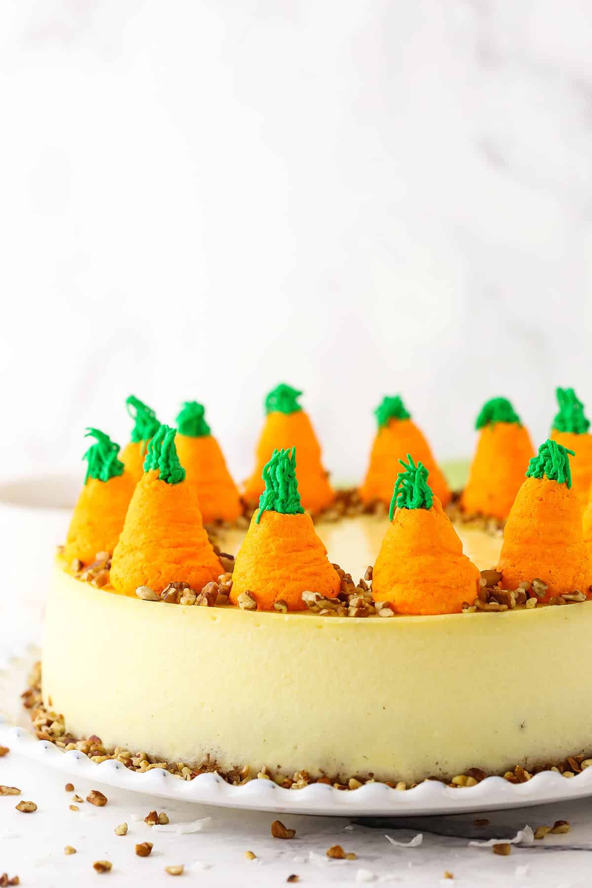 Side view of a full Carrot Cake Cheesecake topped with chopped pecans and icing shaped like carrots on a white platter