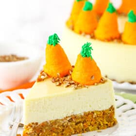 Side view of a slice of Carrot Cake Cheesecake topped with chopped pecans and icing shaped like carrots on a white platter with a fork