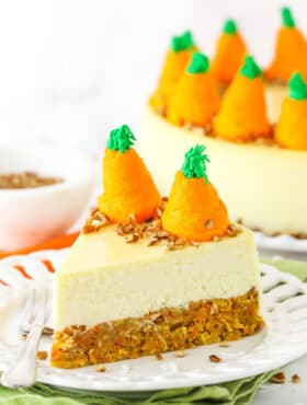 Side view of a slice of Carrot Cake Cheesecake topped with chopped pecans and icing shaped like carrots on a white platter with a fork