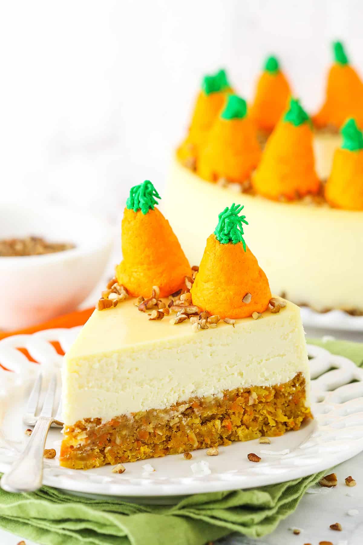 Side view of a slice of Carrot Cake Cheesecake topped with chopped pecans and icing shaped like carrots on a white platter with a fork