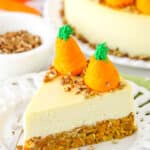 Side view of a slice of Carrot Cake Cheesecake topped with chopped pecans and icing shaped like carrots on a white platter with a fork