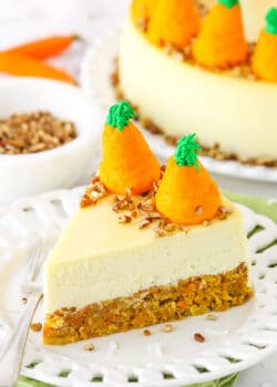 Side view of a slice of Carrot Cake Cheesecake topped with chopped pecans and icing shaped like carrots on a white platter with a fork