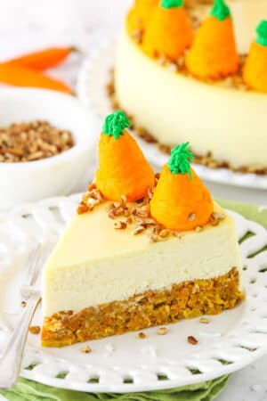 Side view of a slice of Carrot Cake Cheesecake topped with chopped pecans and icing shaped like carrots on a white platter with a fork