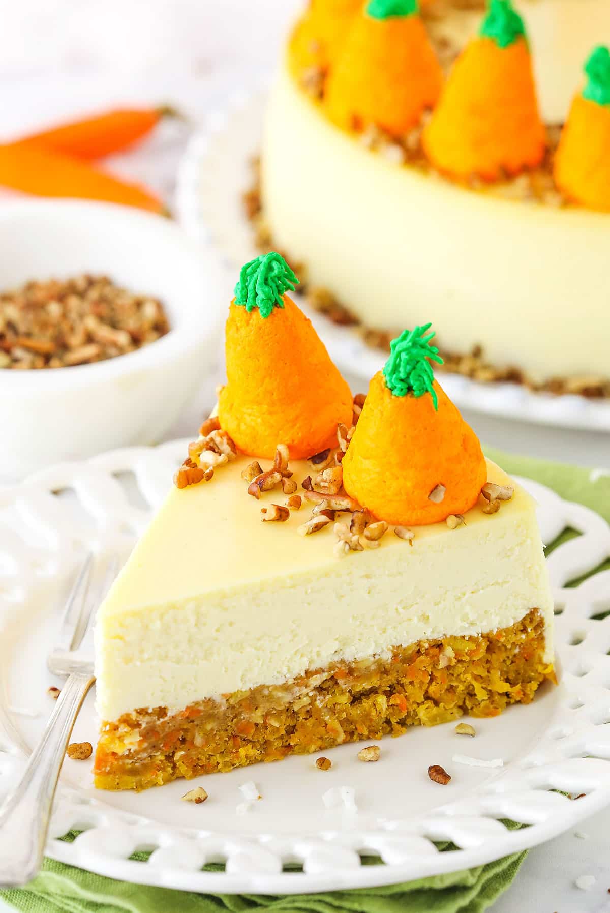 Side view of a slice of Carrot Cake Cheesecake topped with chopped pecans and icing shaped like carrots on a white platter with a fork