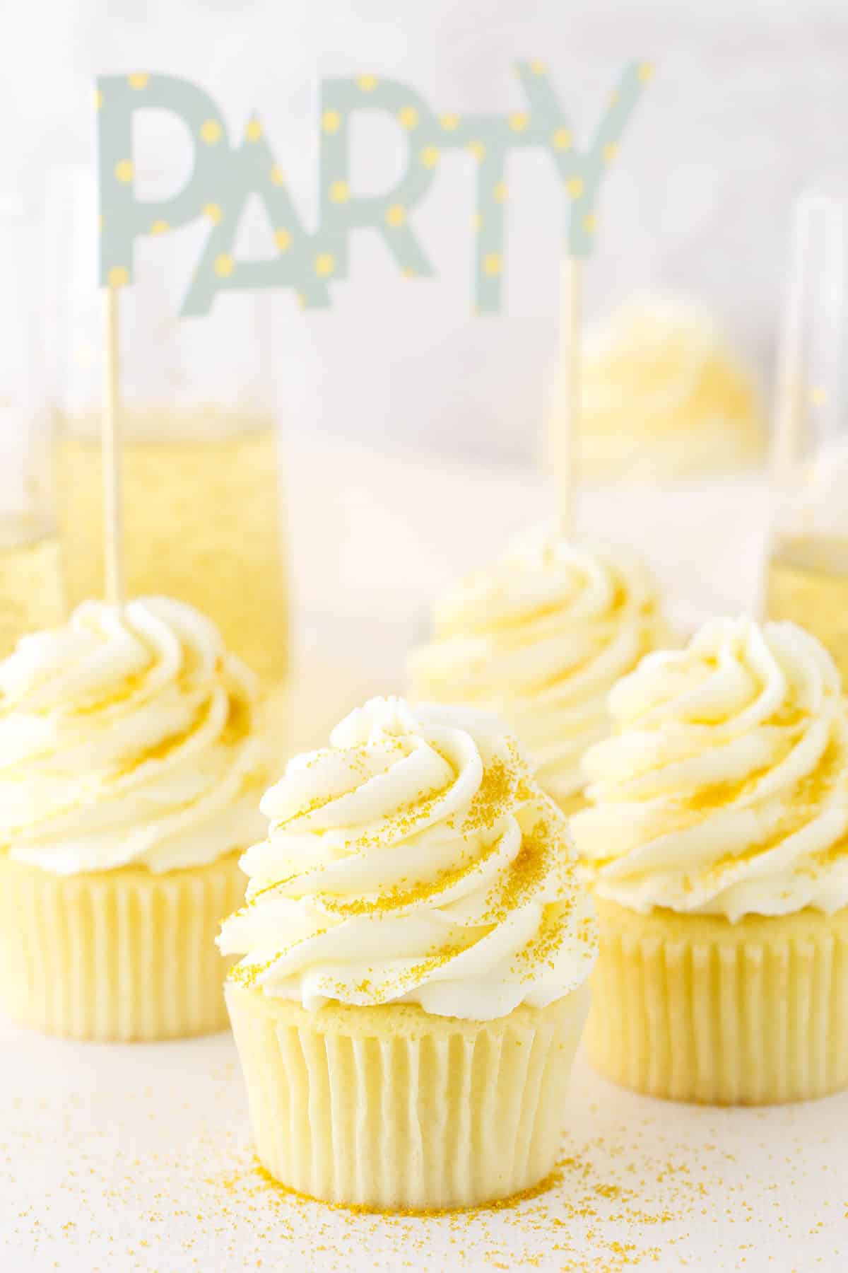 Four Champagne Cupcakes decorated with buttercream frosting on a white table top with a filled champagne glass in the background