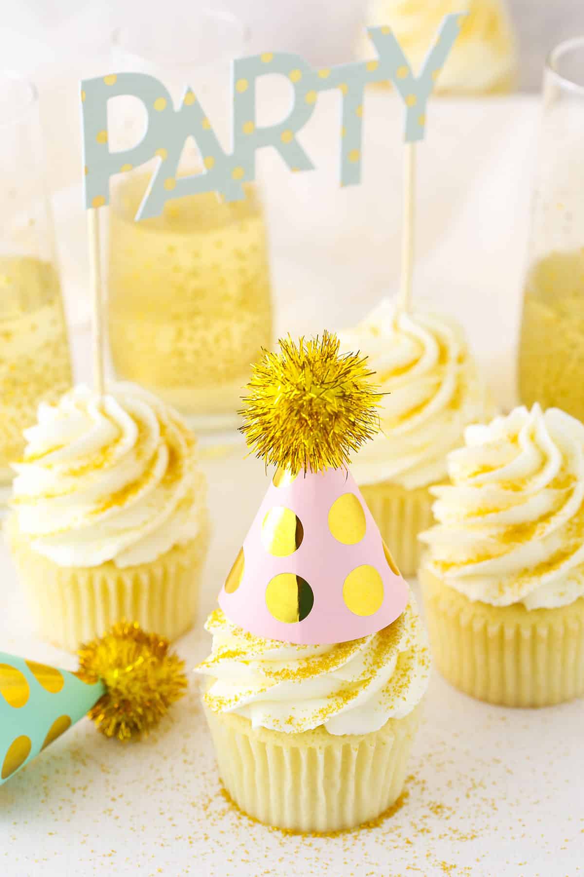 Four Champagne Cupcakes decorated with buttercream frosting on a white table top with a filled champagne glasses in the background