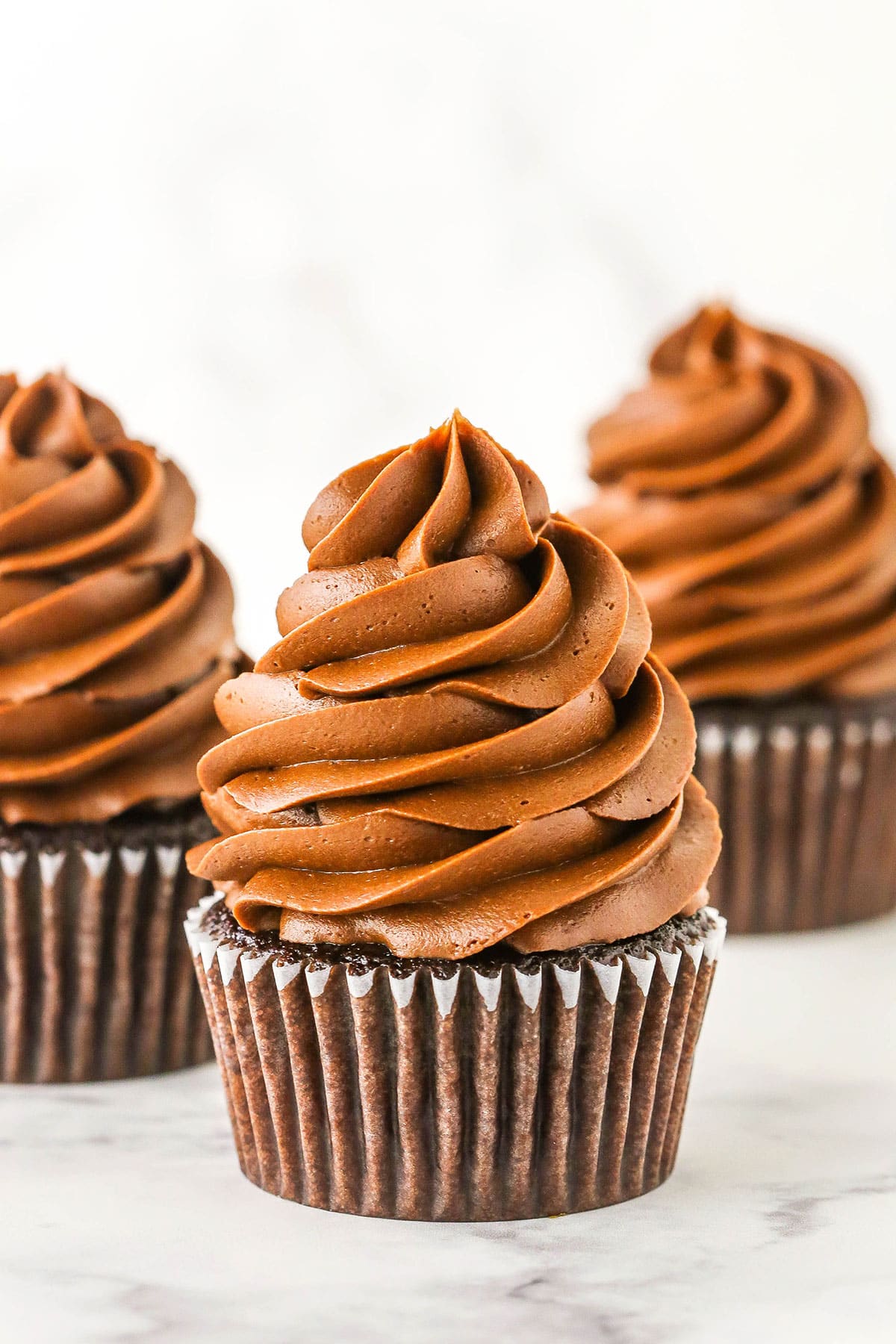 Three chocolate cupcakes topped with Chocolate Buttercream Frosting on a white marble table