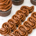 Two rows of Chocolate Buttercream Frosting piped onto a white marble table with three chocolate cupcakes topped with Chocolate Buttercream Frosting in the background