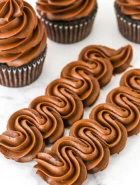 Two rows of Chocolate Buttercream Frosting piped onto a white marble table with three chocolate cupcakes topped with Chocolate Buttercream Frosting in the background