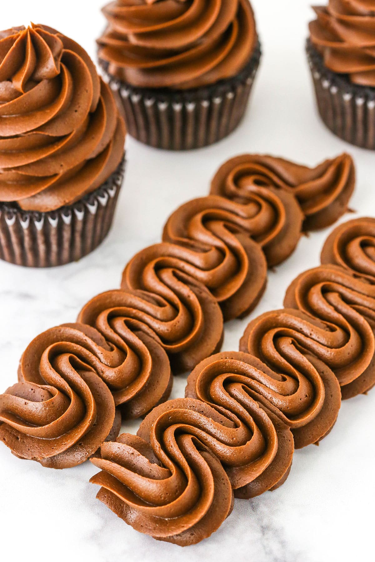 Two rows of Chocolate Buttercream Frosting piped onto a white marble table with three chocolate cupcakes topped with Chocolate Buttercream Frosting in the background