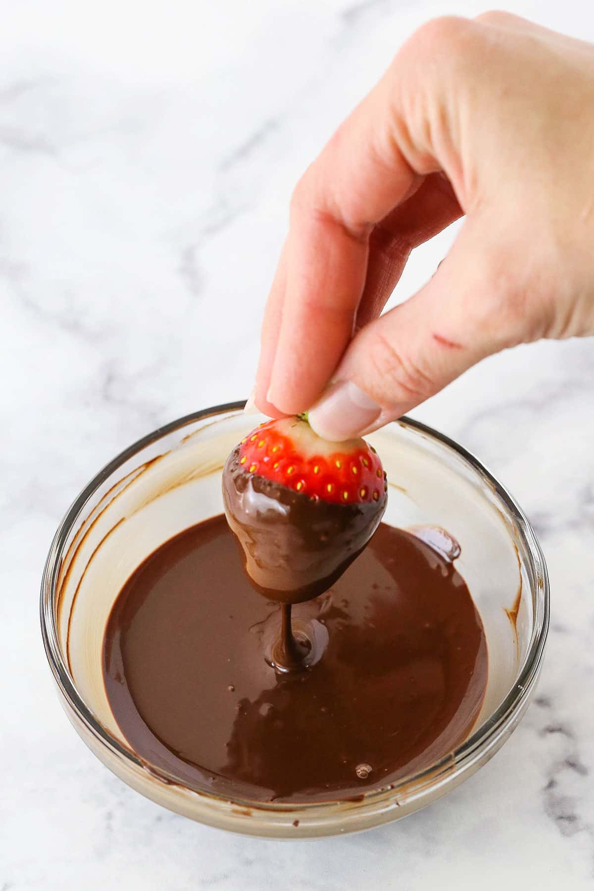 A strawberry being dipped by hand into a glass bowl with melted chocolate