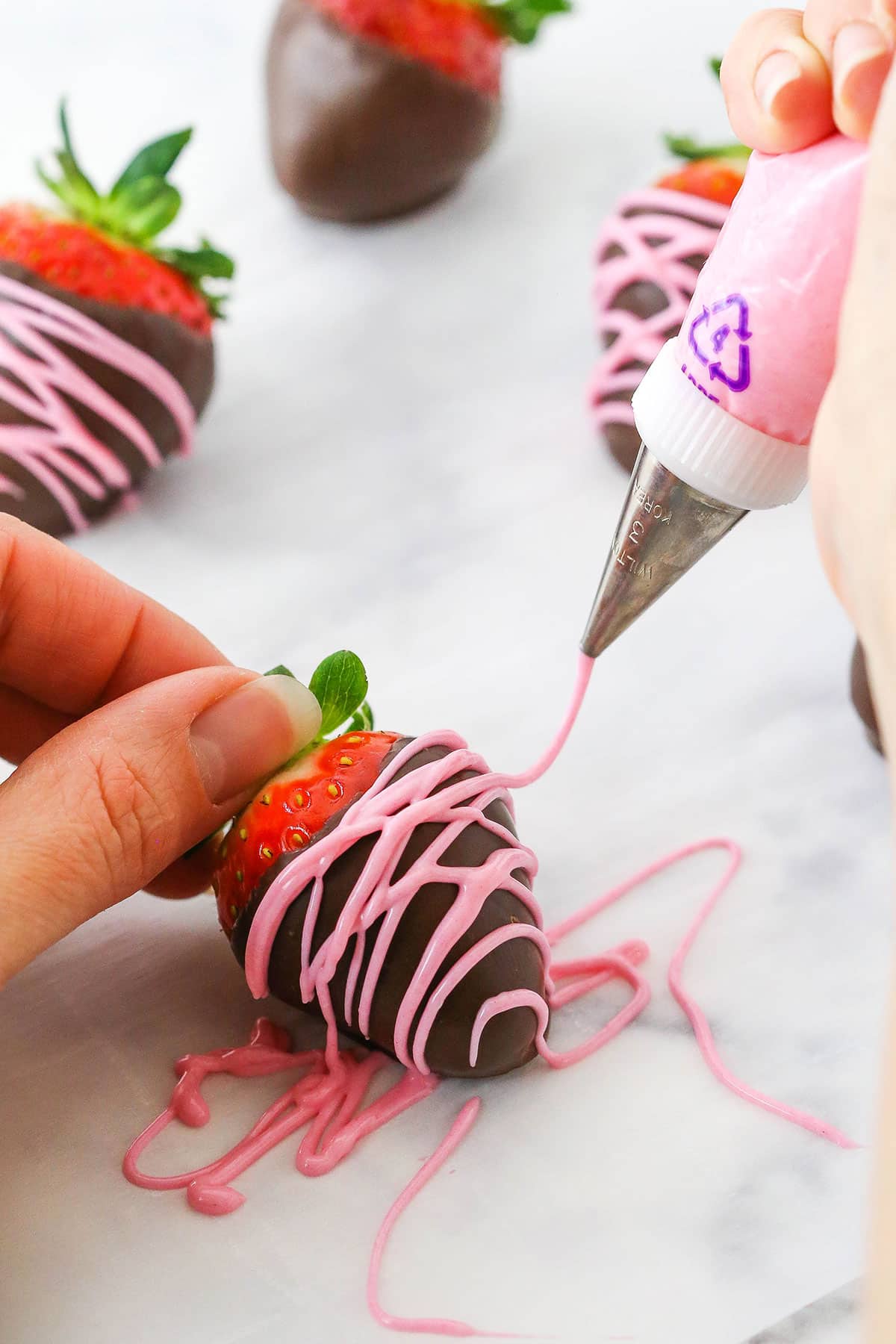 A Chocolate Covered Strawberry being decorated with pink frosting using a piping bag and small piping tip