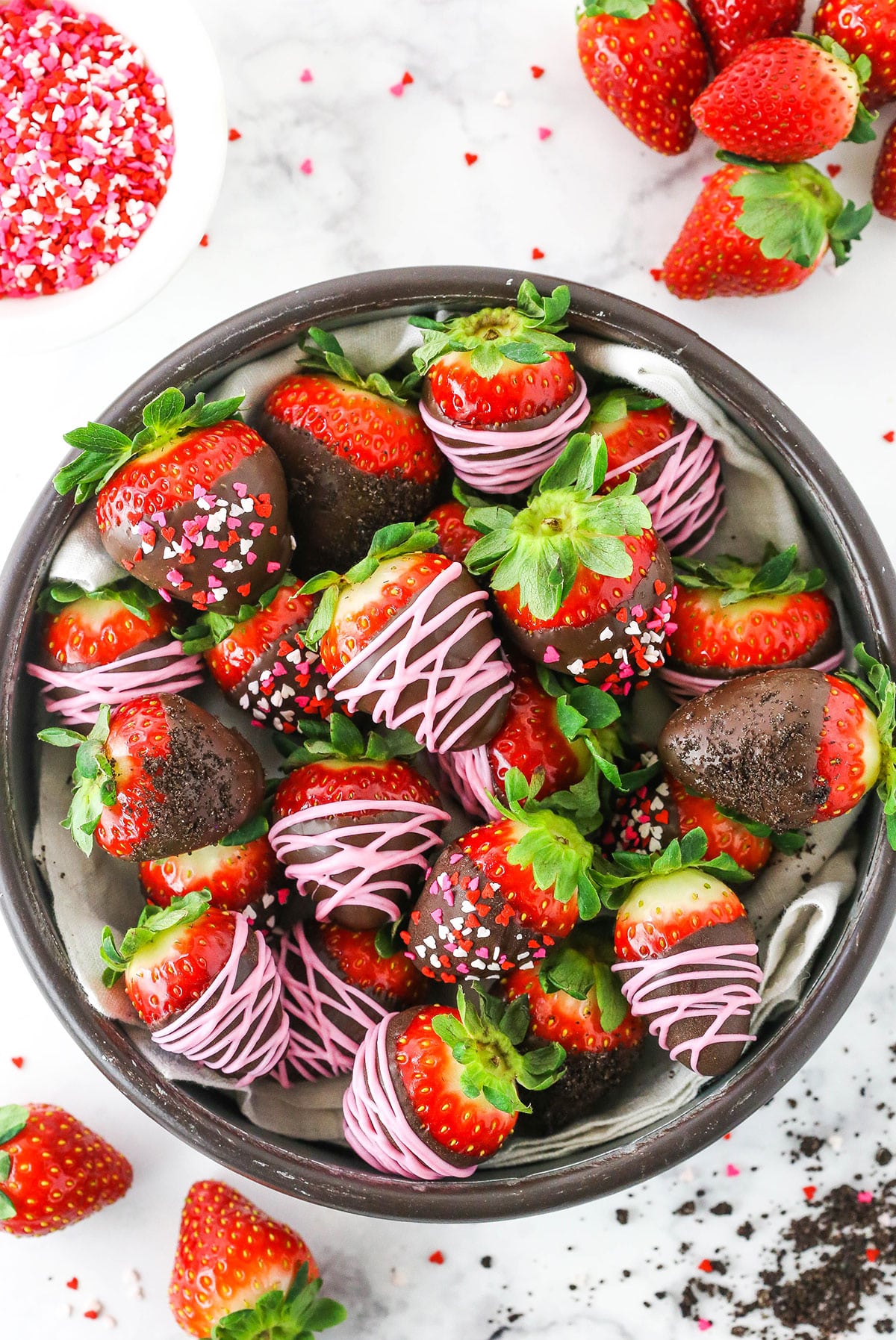 Overhead view of Chocolate Covered Strawberries decorated with pink frosting or red, white and pink sprinkles stacked in a glass bowl on a white table top