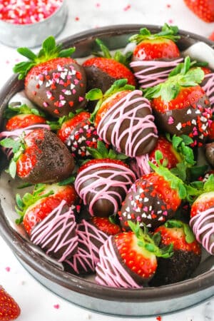 Chocolate Covered Strawberries decorated with pink frosting or red, white and pink sprinkles stacked in a glass bowl on a white table top