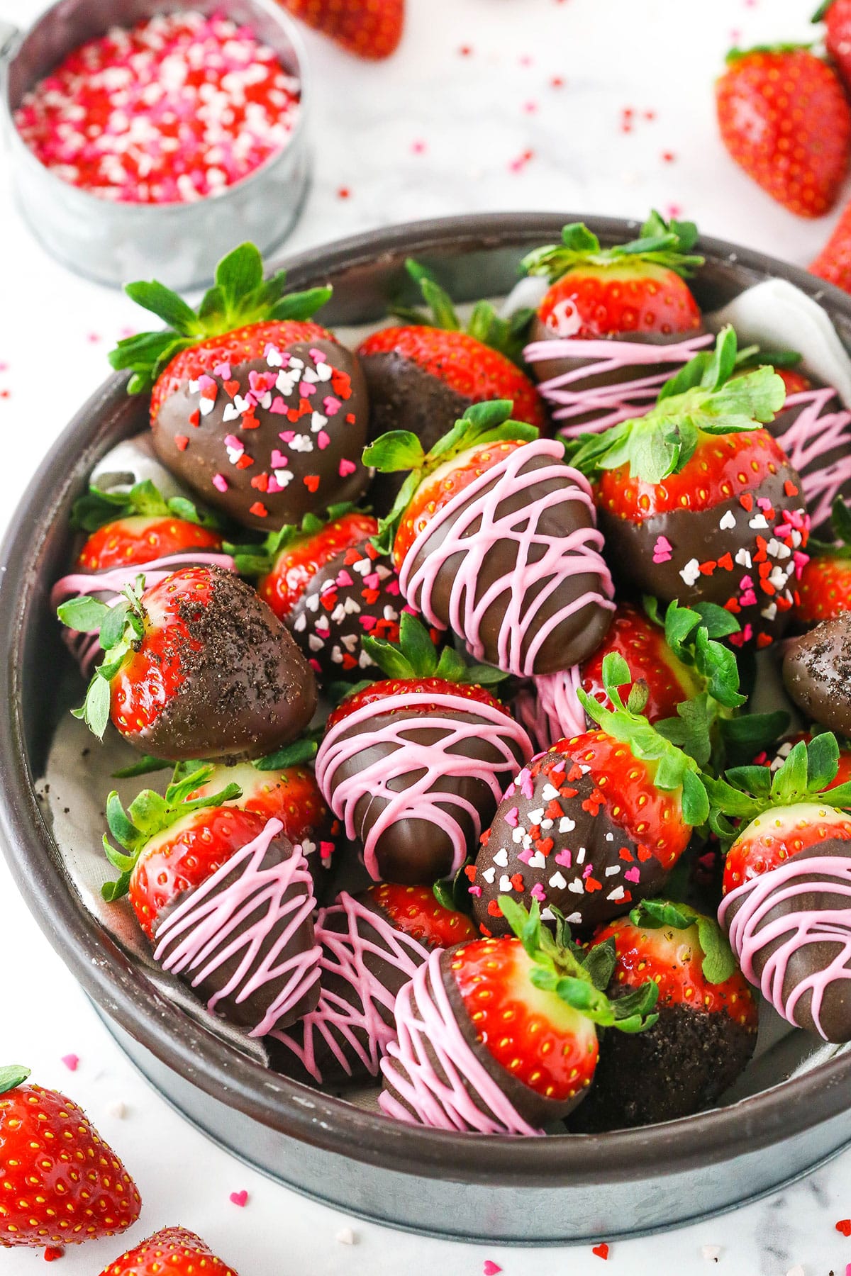 Chocolate Covered Strawberries decorated with pink frosting or red, white and pink sprinkles stacked in a glass bowl on a white table top