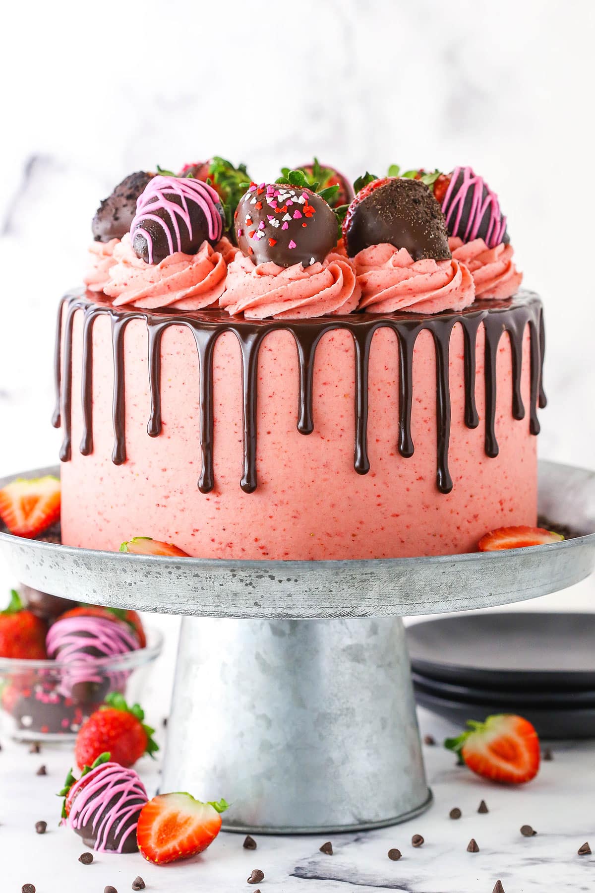 Side view of a full Chocolate Covered Strawberry Layer Cake topped with chocolate drip, pink swirls and chocolate covered strawberries on a metal cake stand