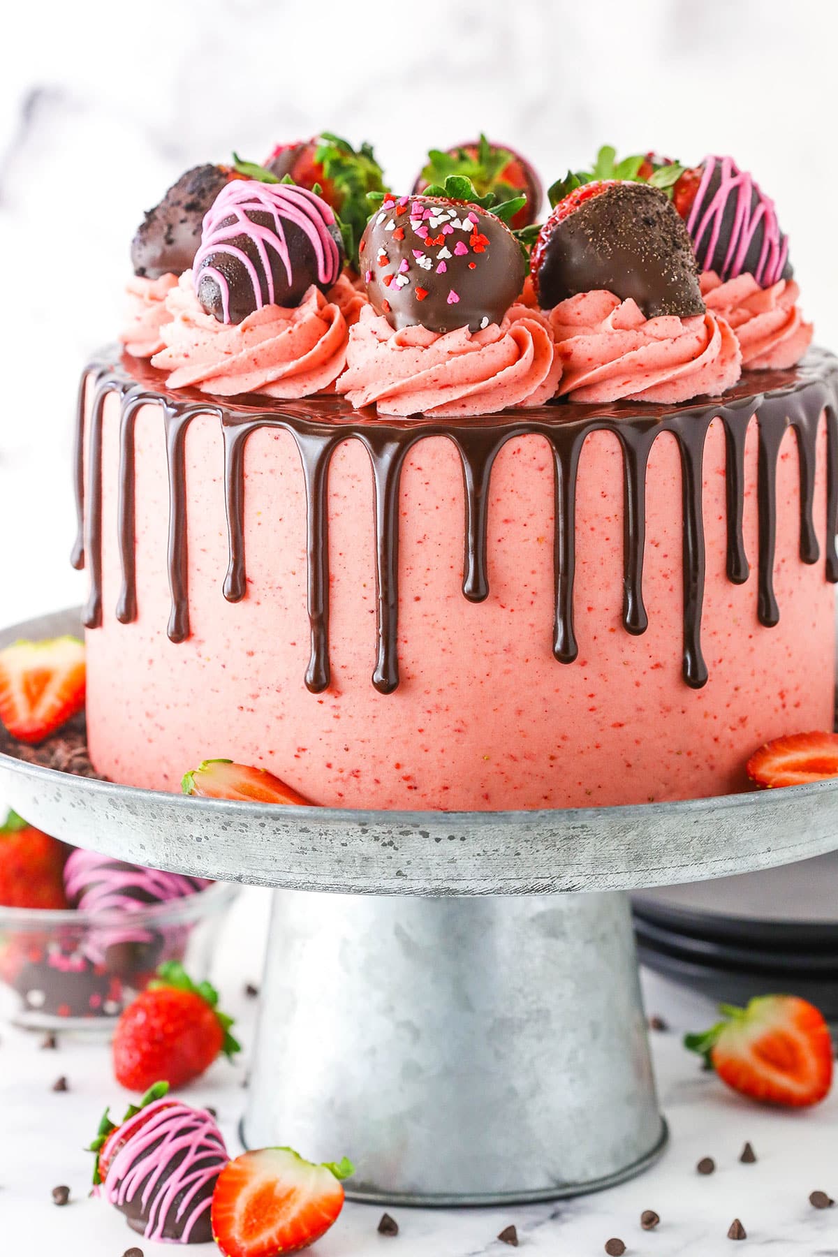 Side view of a full Chocolate Covered Strawberry Layer Cake topped with chocolate drip, pink swirls and chocolate covered strawberries on a metal cake stand