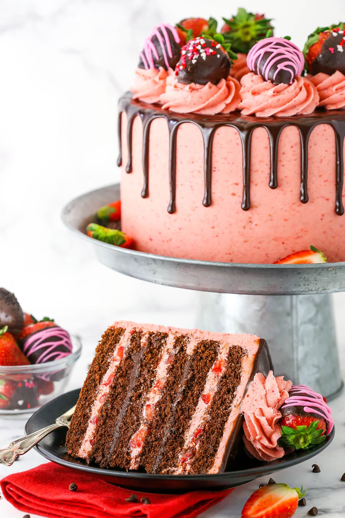 A slice of Chocolate Covered Strawberry Layer Cake with a fork on a black plate with the Chocolate Covered Strawberry Layer Cake on a metal cake stand in the background