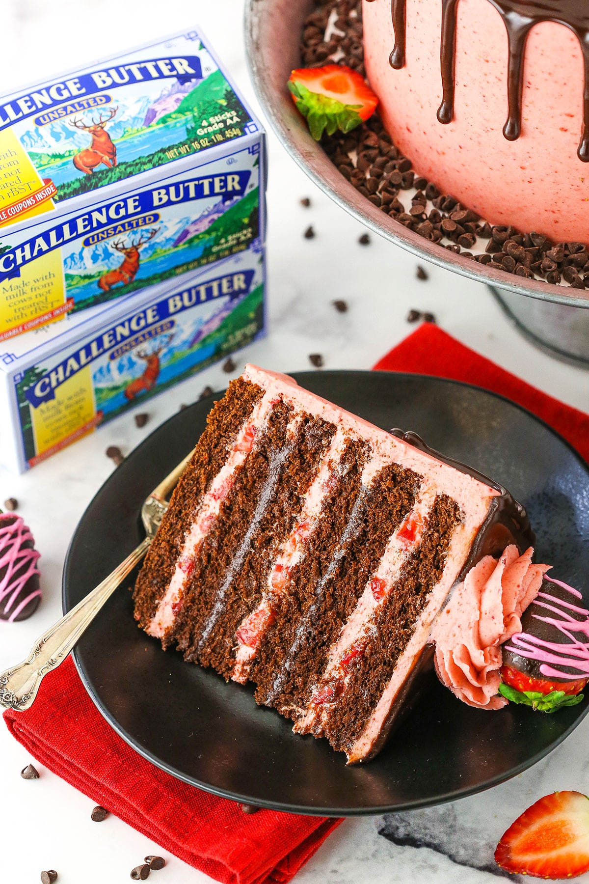 A slice of Chocolate Covered Strawberry Layer Cake on a black plate with a fork and Challenge Butter in the background