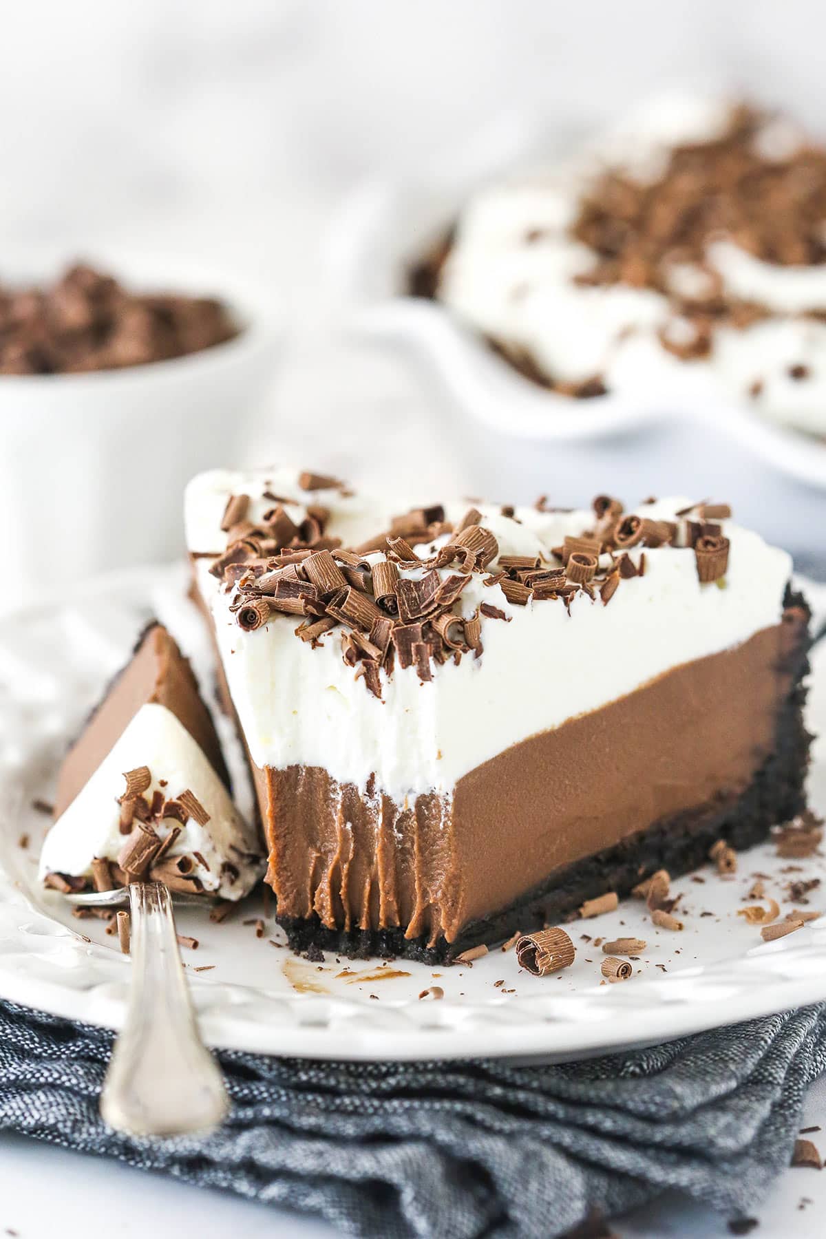 Chocolate Cream Pie is made with Oreo pie crust, chocolate pudding filling, whipped cream, and a smattering of chocolate shavings and curls. Chocolate cream pie on a plate with a fork taking a bite out of it.