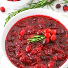 Cranberry sauce in a white bowl surrounded by cranberries and rosemary.