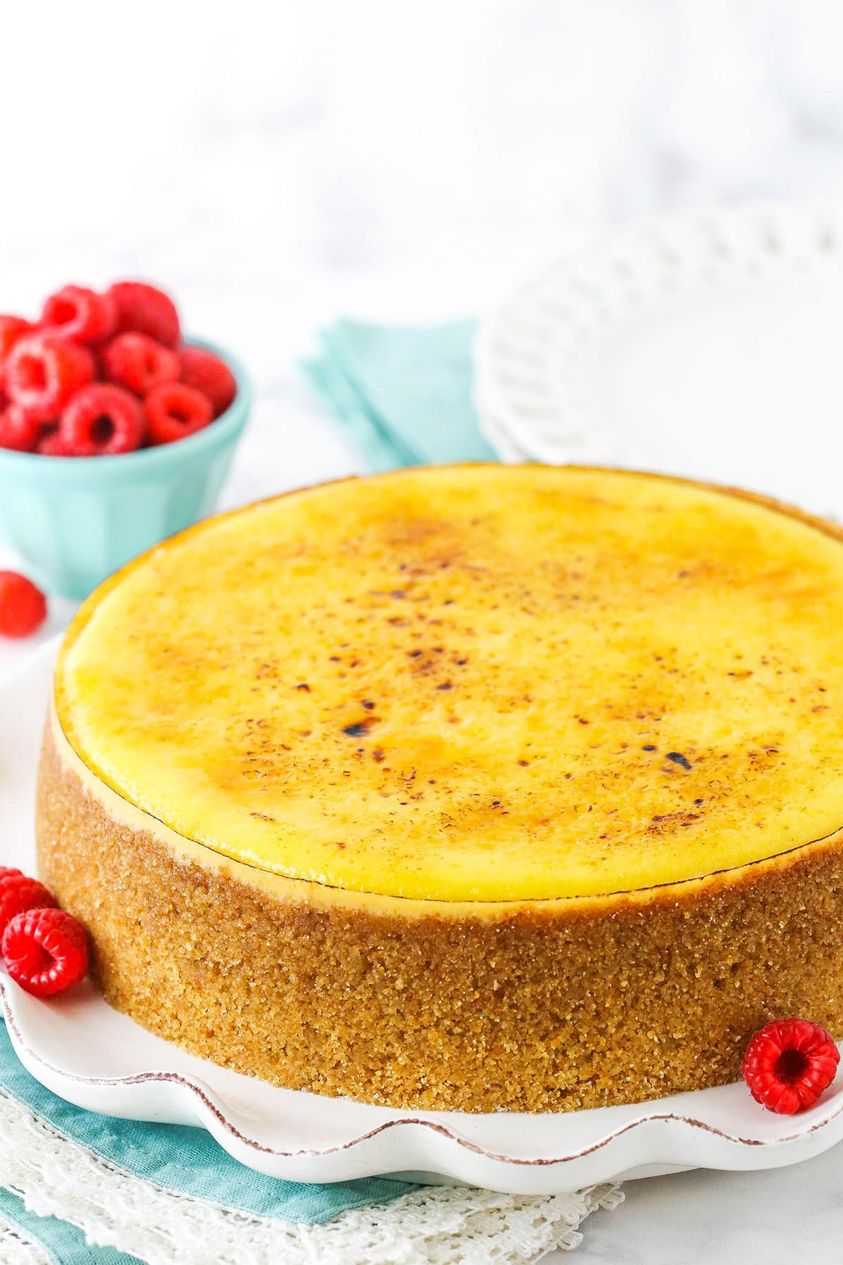 A full Creme Brûlée Cheesecake on a white platter with raspberries in the background