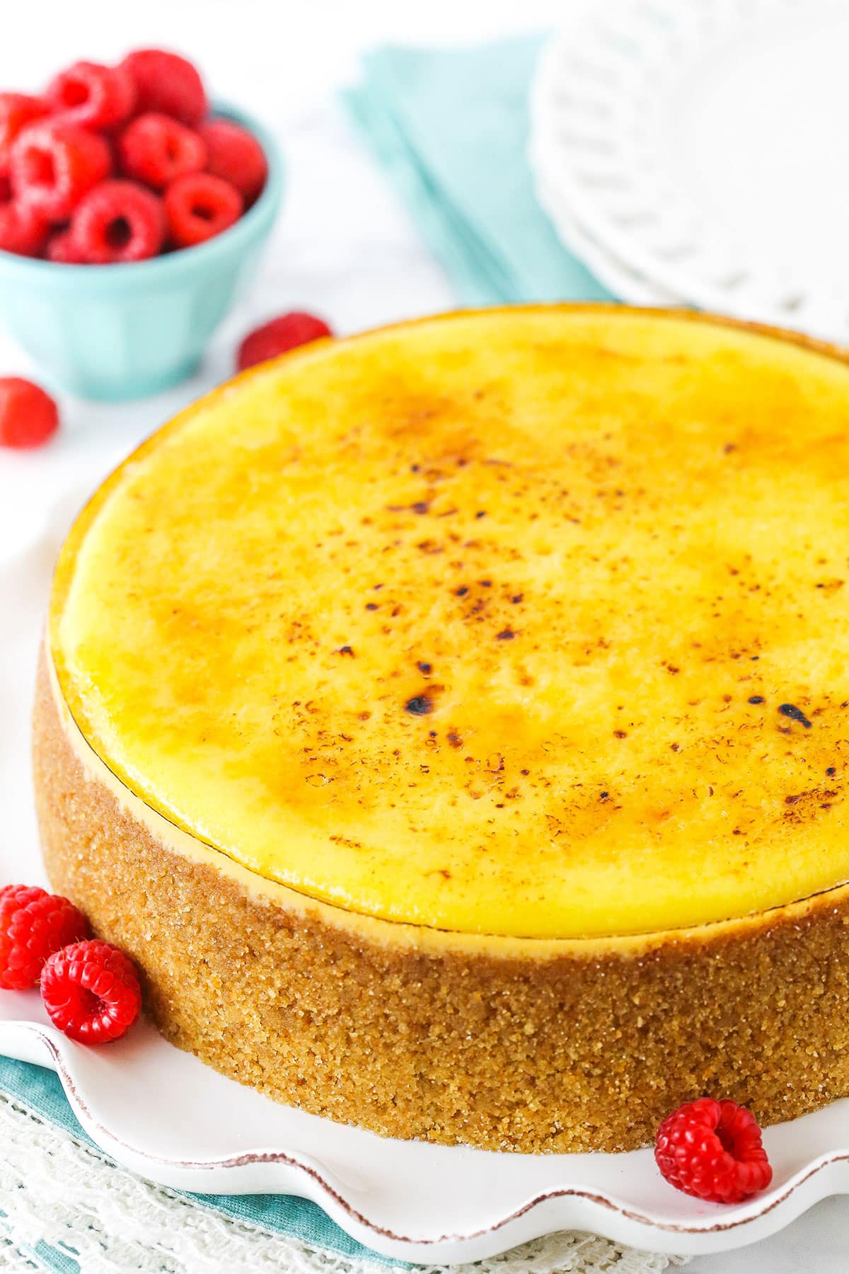 A full Creme Brûlée Cheesecake on a white platter with raspberries in the background
