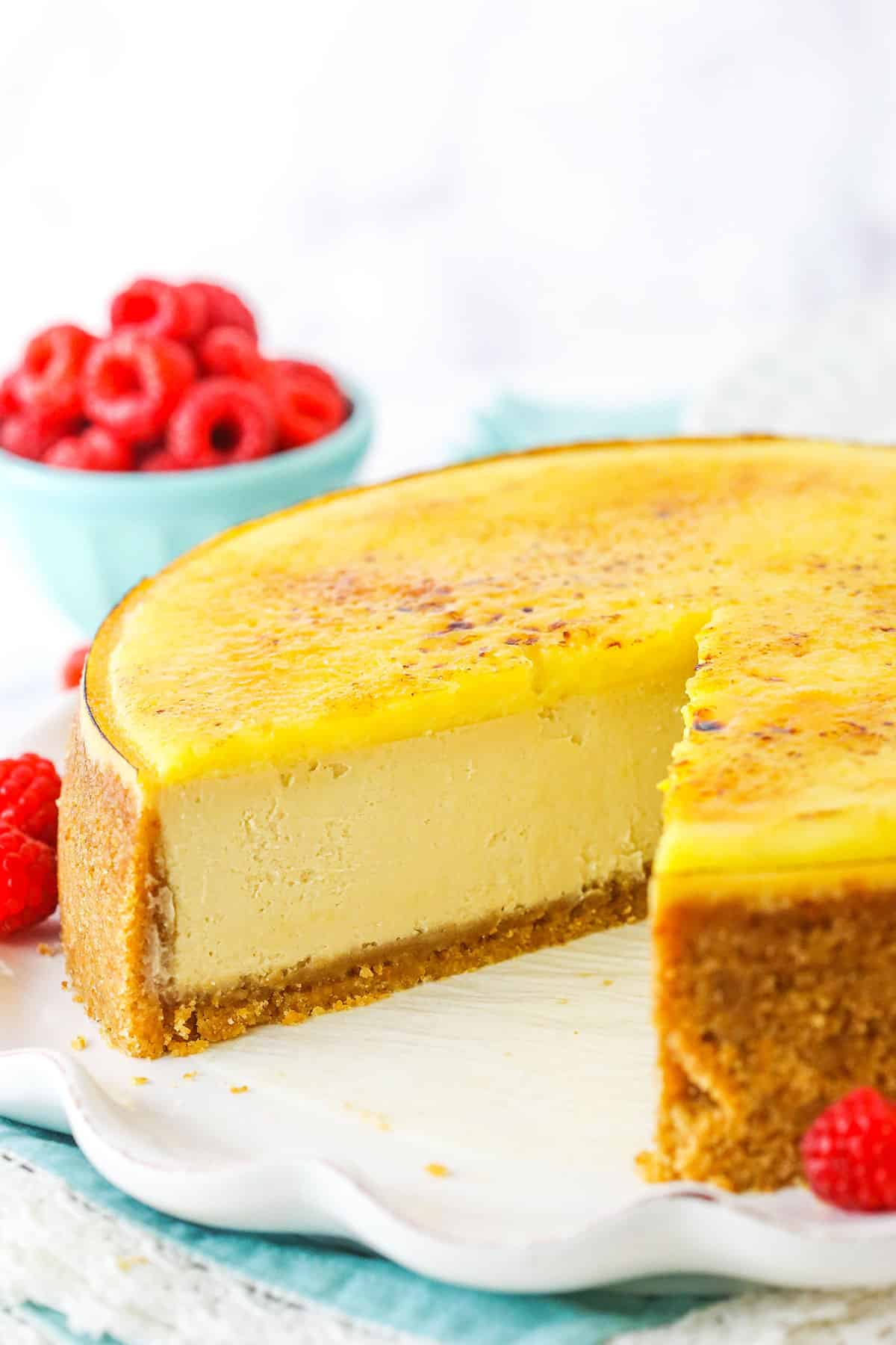 Side view of a Creme Brûlée Cheesecake with a slice removed on a white platter with raspberries in the background