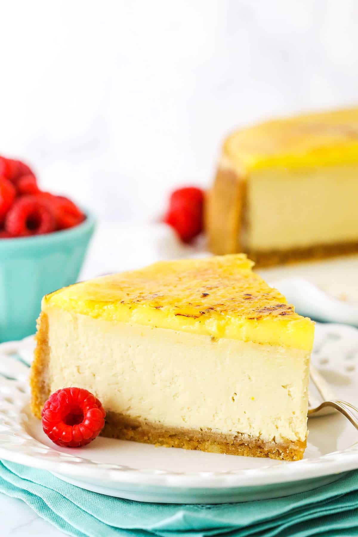 A slice of Creme Brûlée Cheesecake on a white plate with a fork and a raspberry