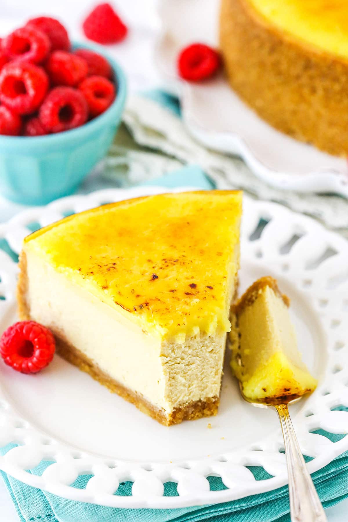 A slice of Creme Brûlée Cheesecake with a bite removed on a white plate with a fork and a raspberry