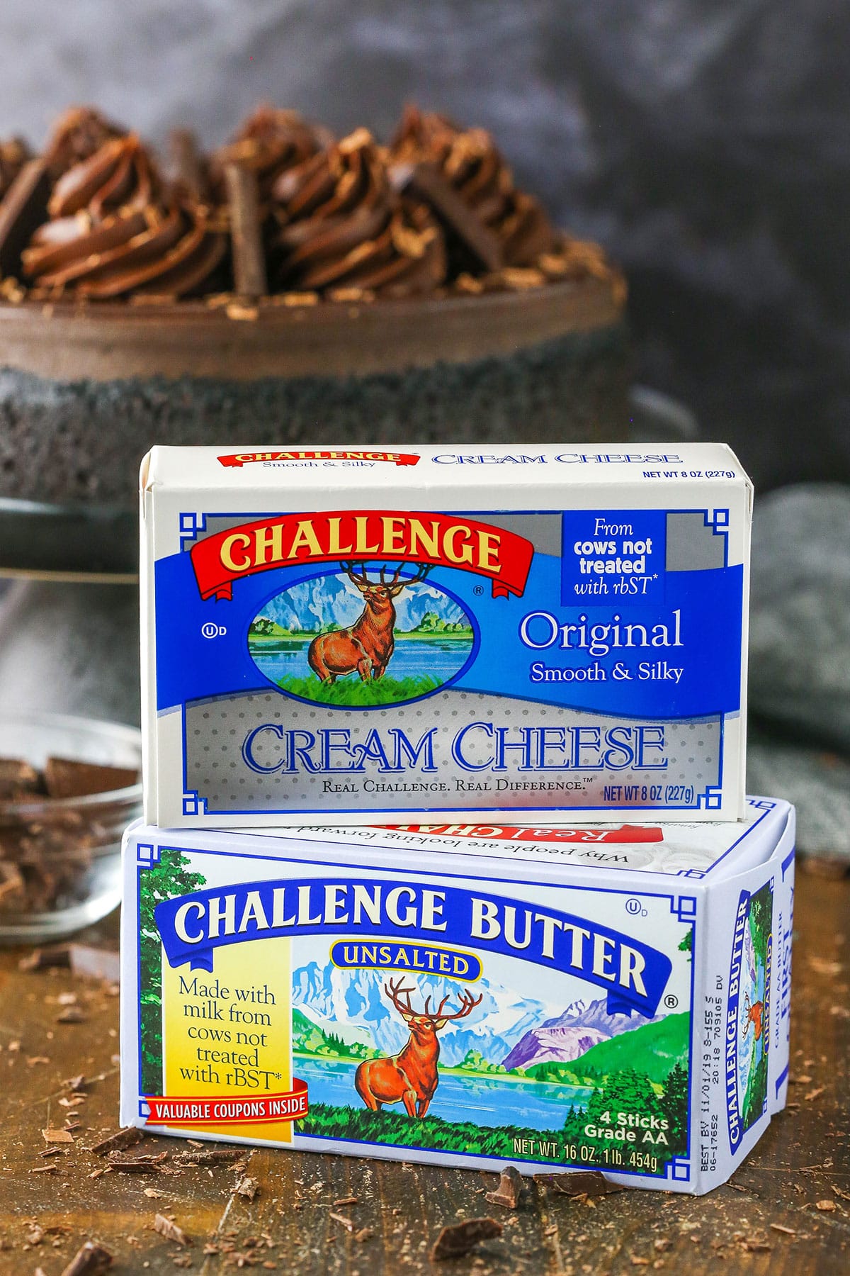 Two packages of Challenge Butter stacked with a Decadent Dark Chocolate Cheesecake in the background