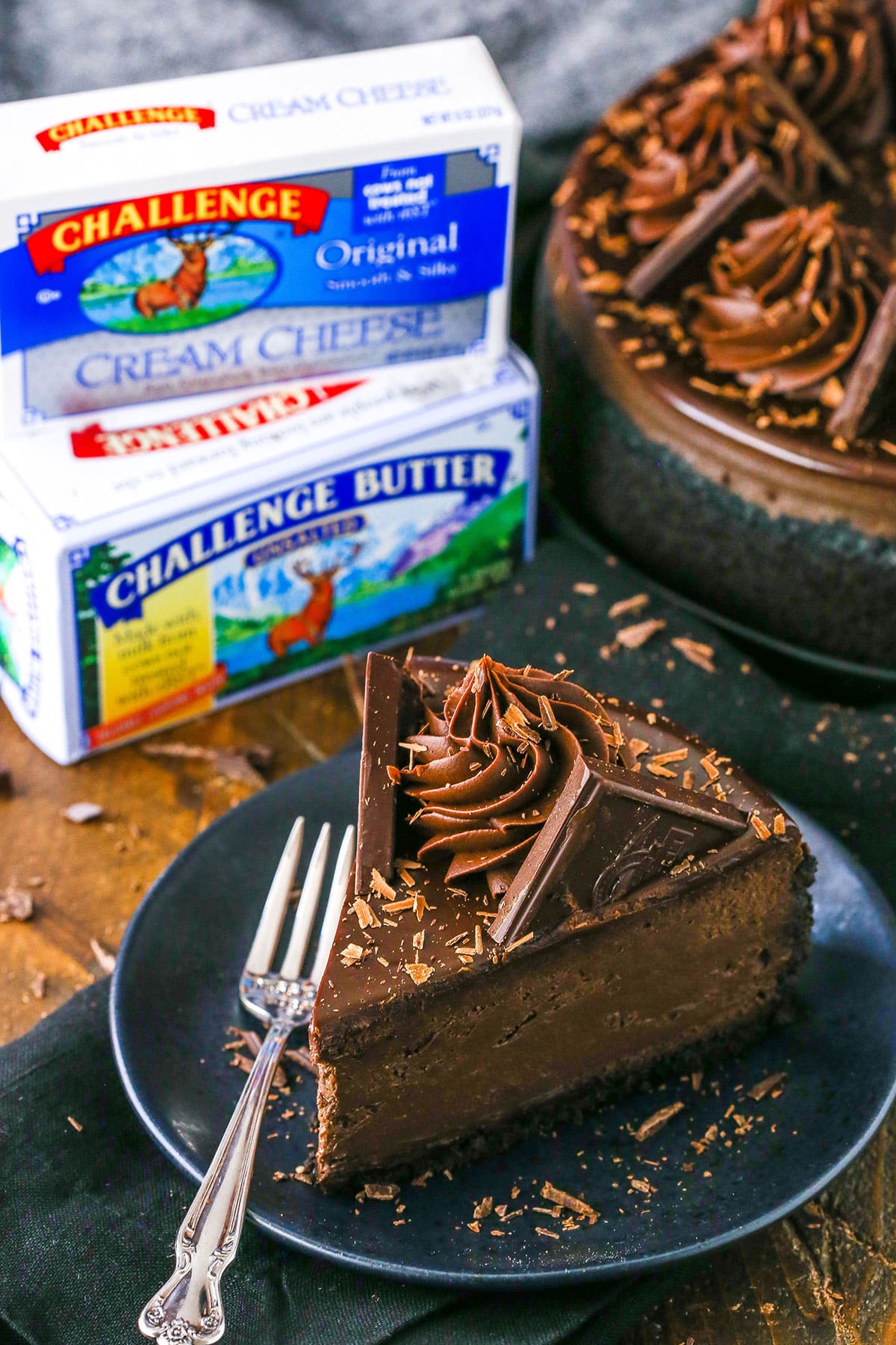 A slice of Decadent Dark Chocolate Cheesecake on a black plate next to a fork with two packages of Challenge Butter in the background