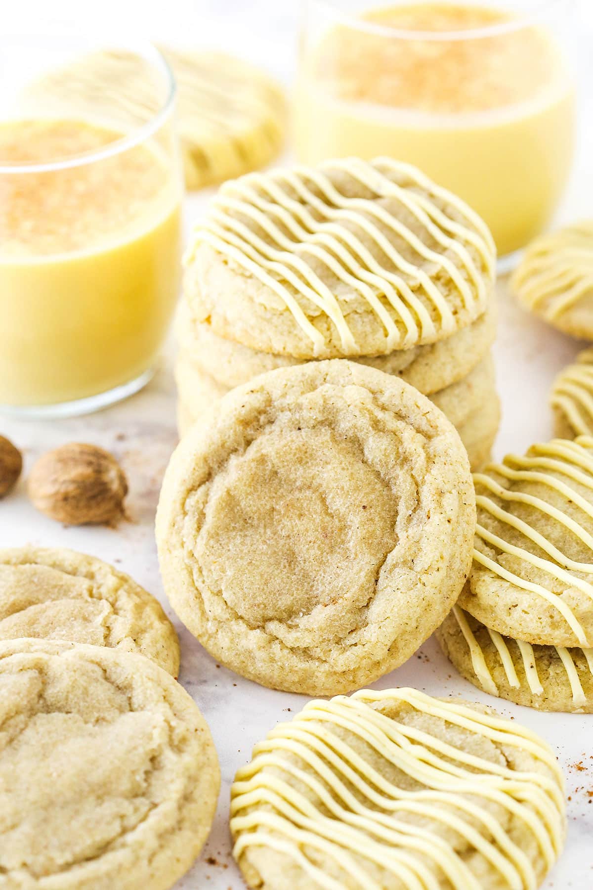 Eggnog Cookies topped with frosting stacked and two glasses of eggnog in the background