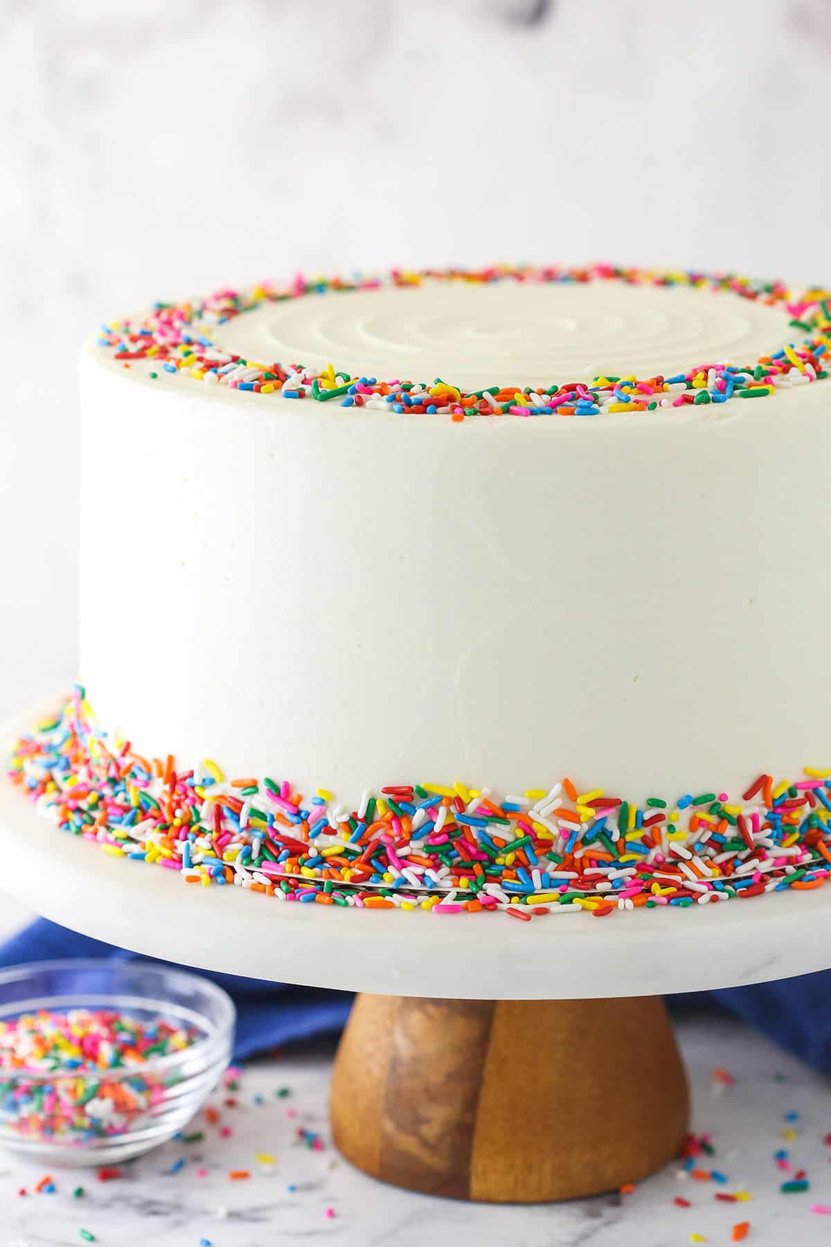 Funfetti layer cake on a cake stand near a bowl of rainbow sprinkles.