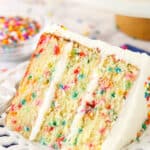 A slice of Funfetti layer cake on a plate surrounded by rainbow sprinkles.