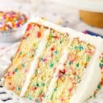 A slice of Funfetti layer cake on a plate surrounded by rainbow sprinkles.