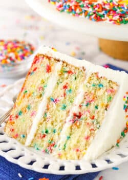 A slice of Funfetti layer cake on a plate surrounded by rainbow sprinkles.