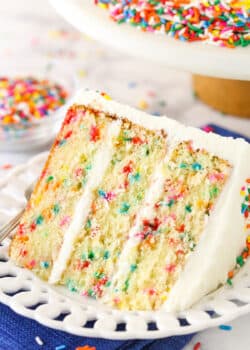 A slice of Funfetti layer cake on a plate surrounded by rainbow sprinkles.