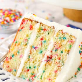 A slice of Funfetti layer cake on a plate surrounded by rainbow sprinkles.