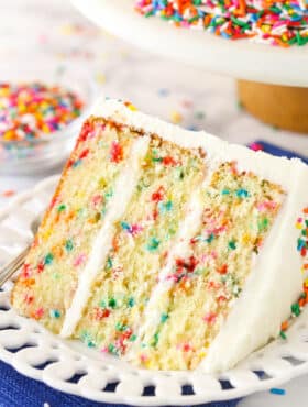 A slice of Funfetti layer cake on a plate surrounded by rainbow sprinkles.