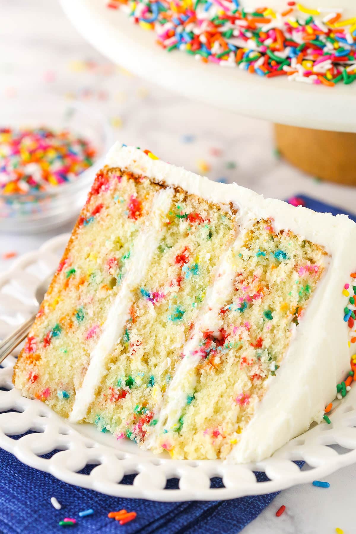 A slice of Funfetti layer cake on a plate surrounded by rainbow sprinkles.