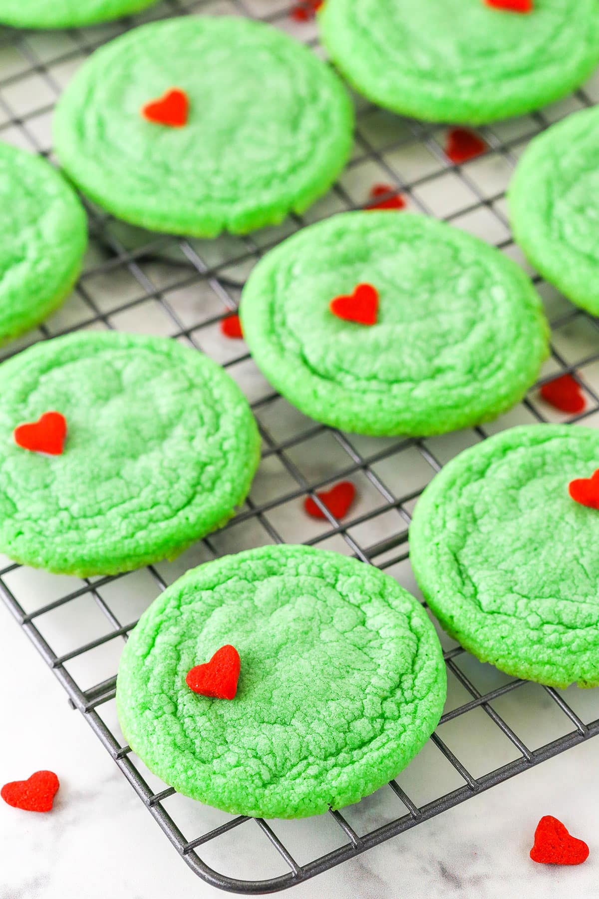 Green Grinch Cookies topped with a small heart shaped sprinkle spread out over a cooling rack on a white table top